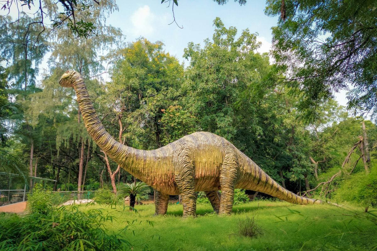 A large dinosaur statue stands amidst lush greenery in a wooded area.