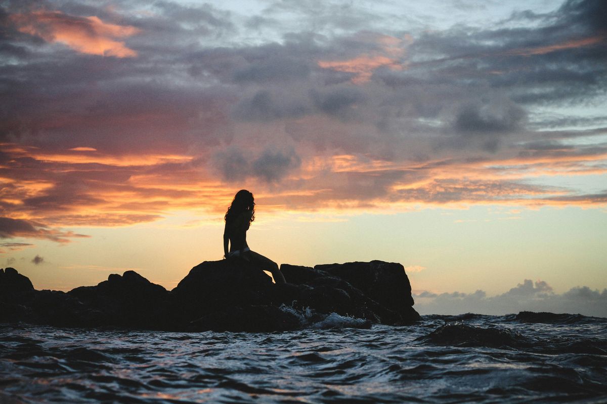 Silhouette of person sitting on rocks against a vibrant sunset sky over the ocean.