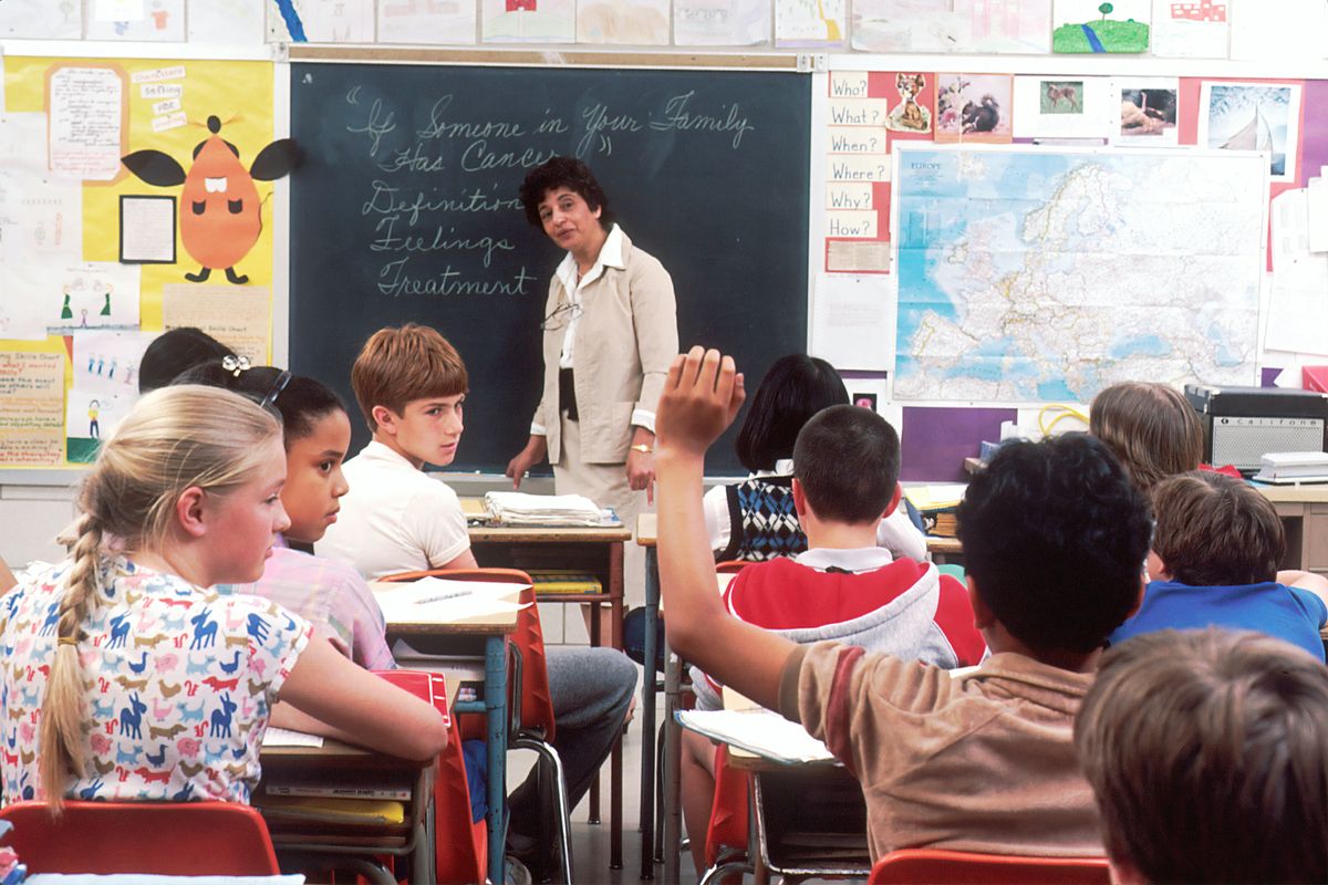 Teacher instructs students in a classroom; one student raises hand to ask a question.