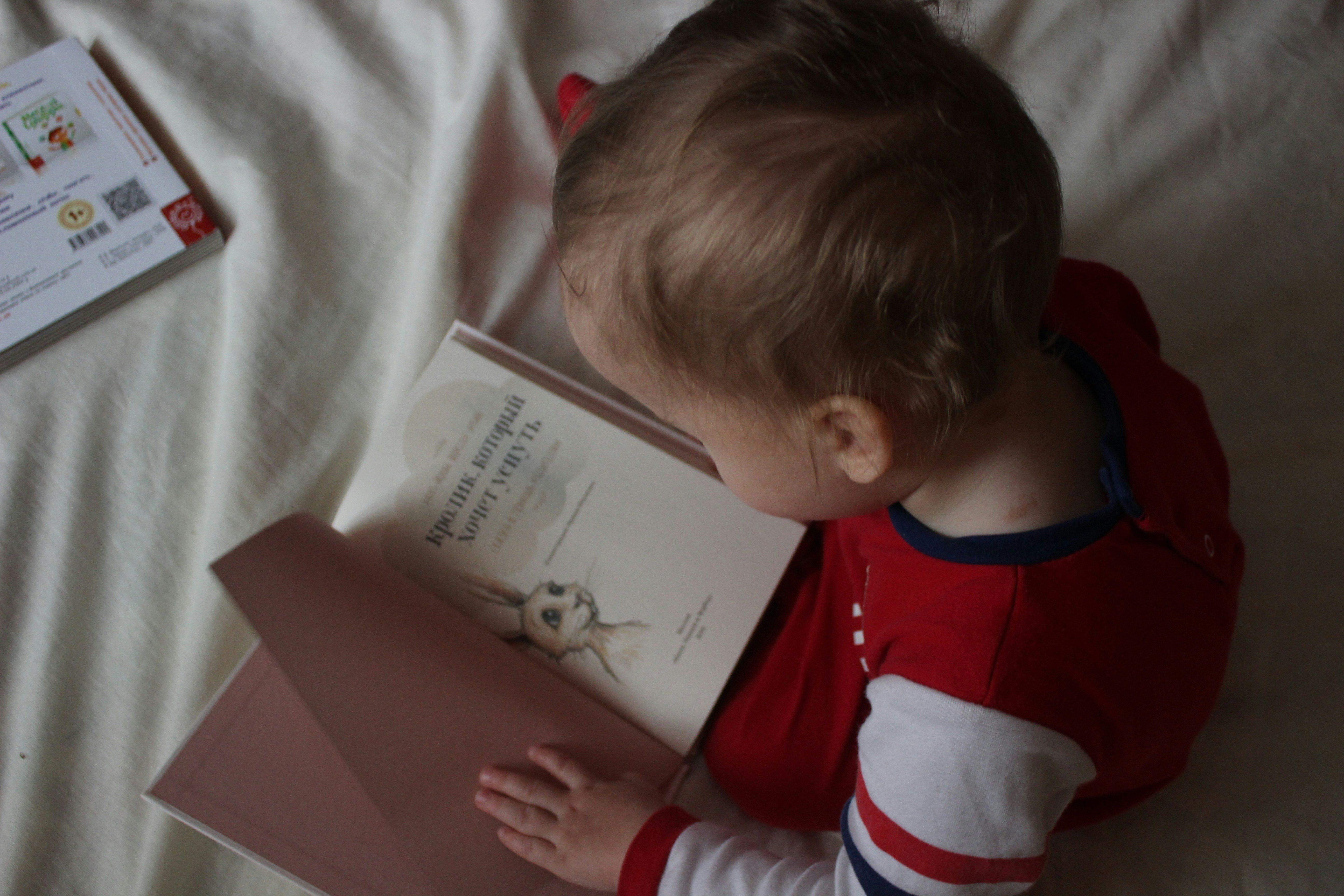 Toddler in red clothes reading a book on a soft white surface.
