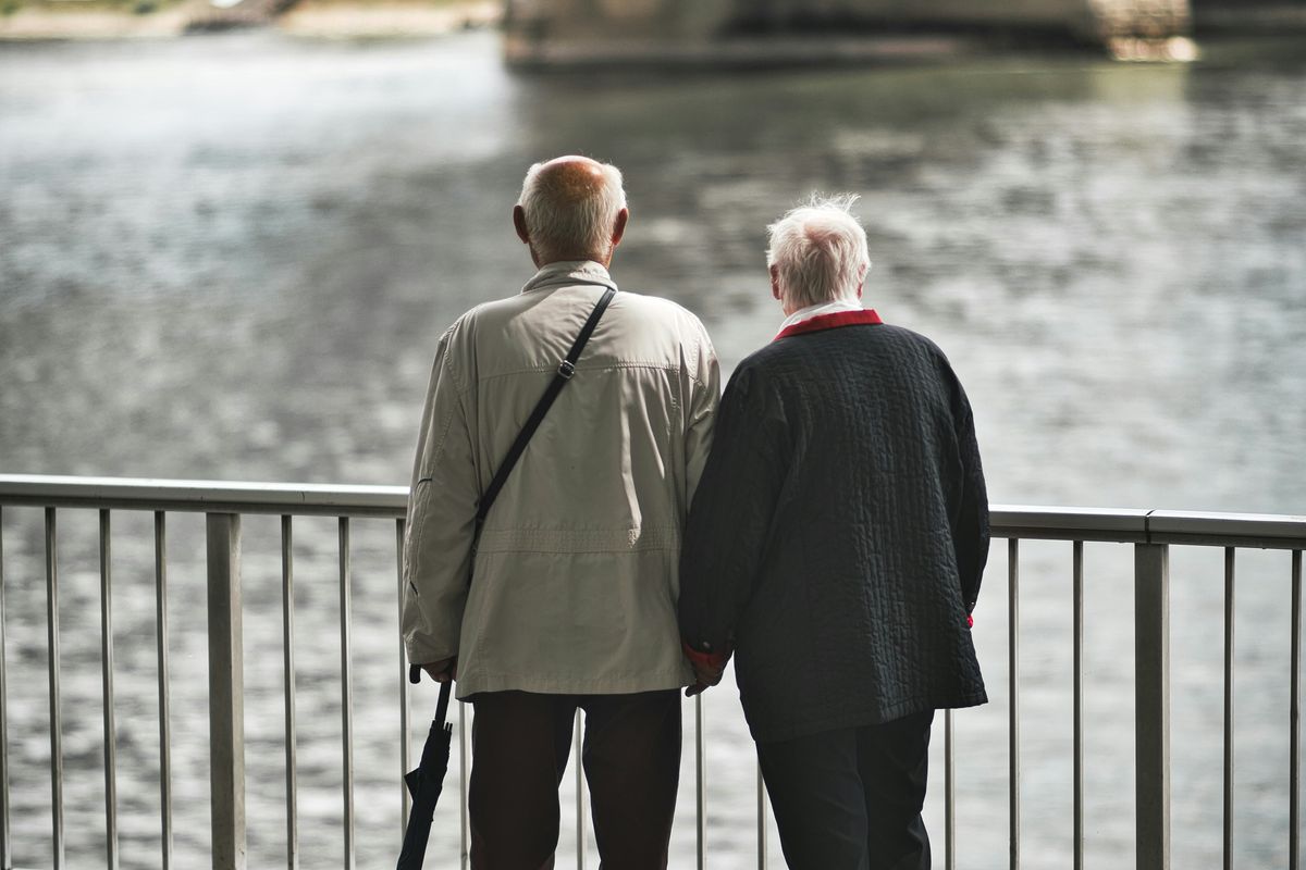 Elderly couple stands by river, holding hands, near a large bridge.