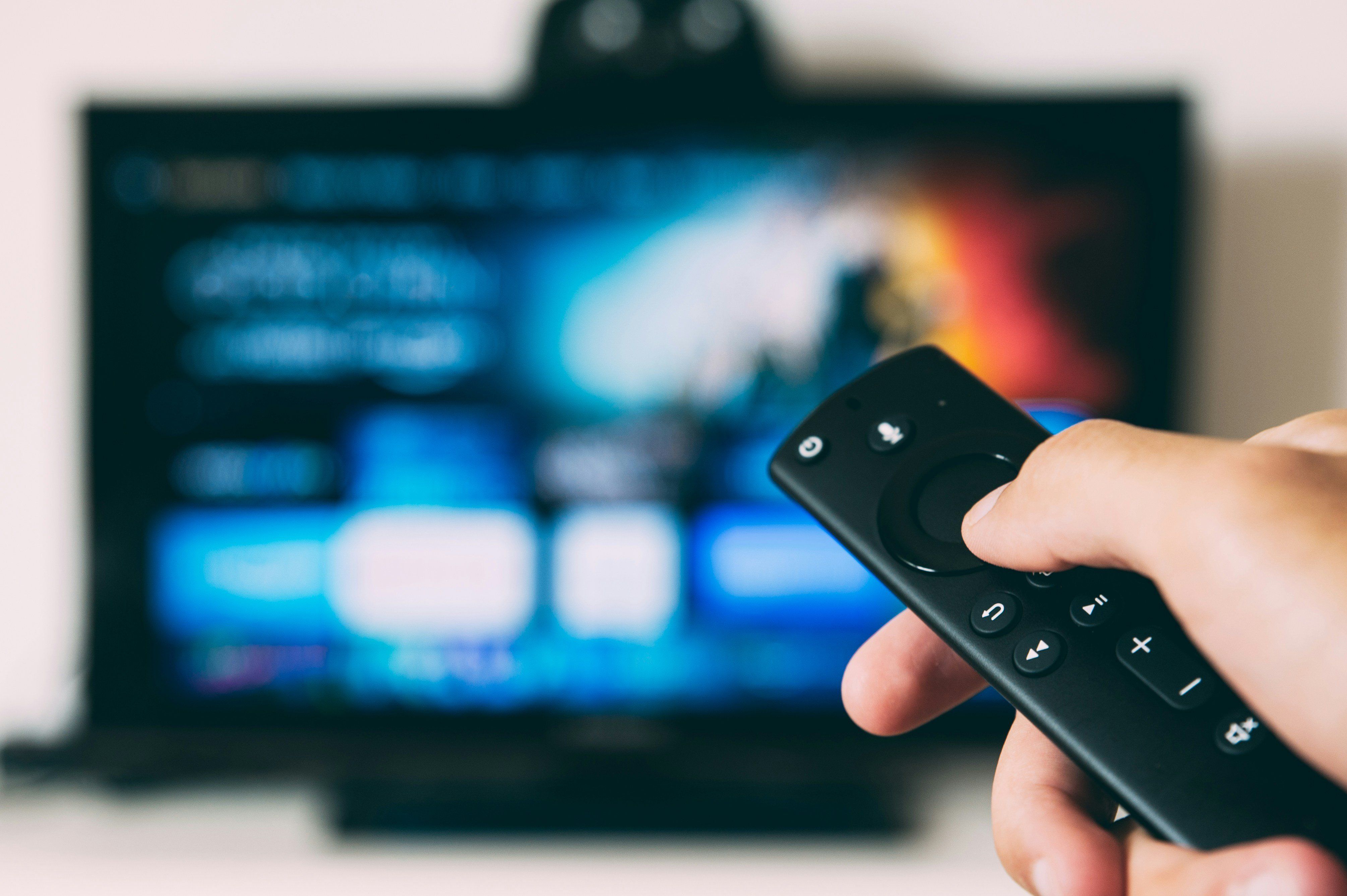 Hand holding a remote control, pointing at a blurred TV screen.