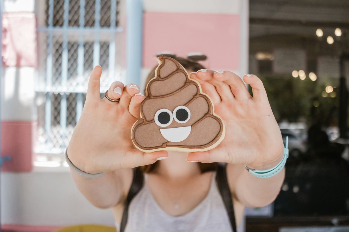 Person holding a poop emoji cookie in front of their face, outdoor setting.