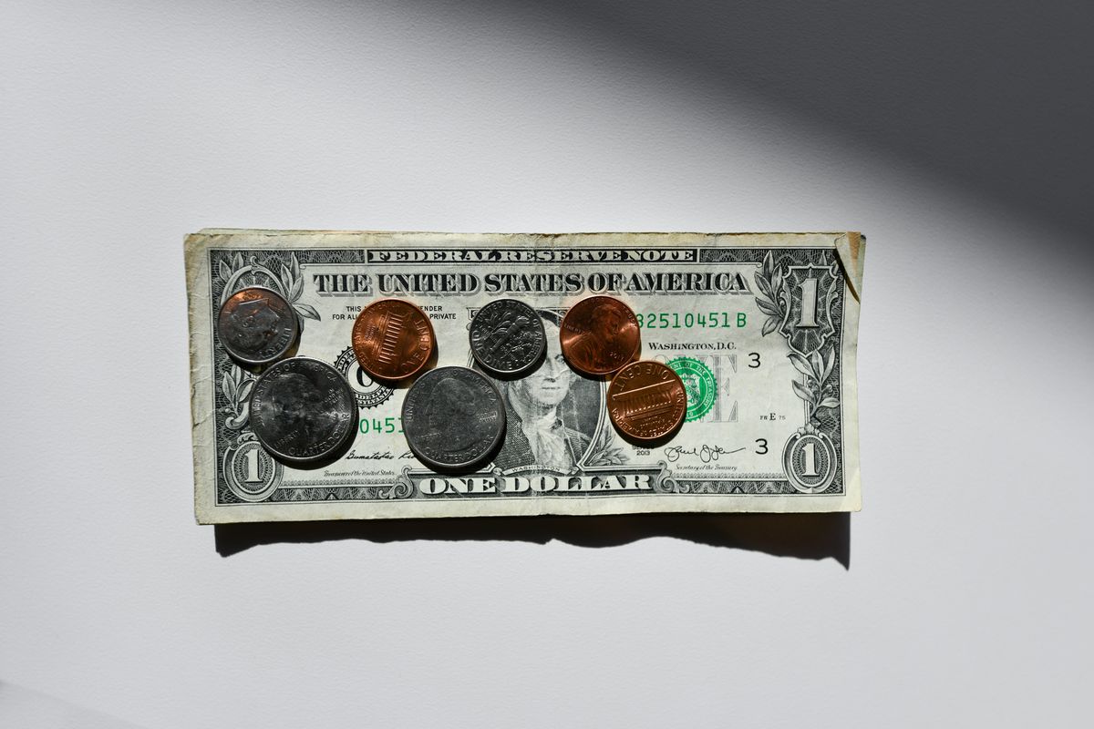 A one-dollar bill with various US coins on top, casting shadows on a white surface.