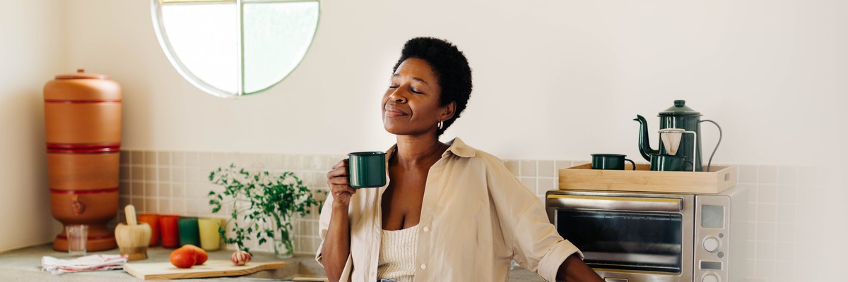 Happy Brazilian woman with afro hair stands in her kitchen, holding an enamel mug of hot coffee, eyes closed, enjoying a peace and quiet moment at home.