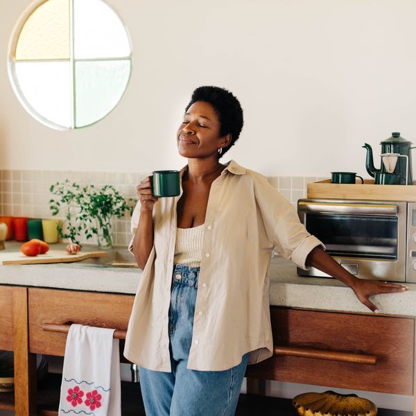 Happy Brazilian woman with afro hair stands in her kitchen, holding an enamel mug of hot coffee, eyes closed, enjoying a peace and quiet moment at home.