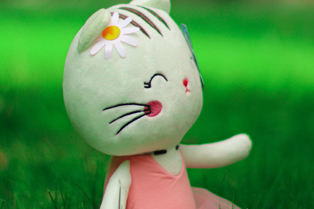 Stuffed cat toy with daisy on its head sits in grass, wearing a pink dress, smiling.