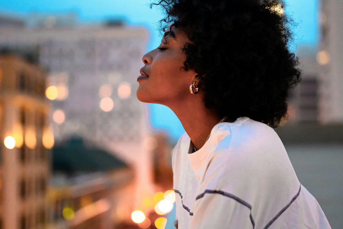 A black woman gazing over a cityscape from a rooftop at dusk.