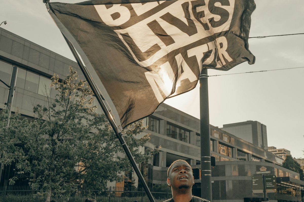 Black guy holding a large "Black Lives Matter" flag at a protest with buildings in the background.