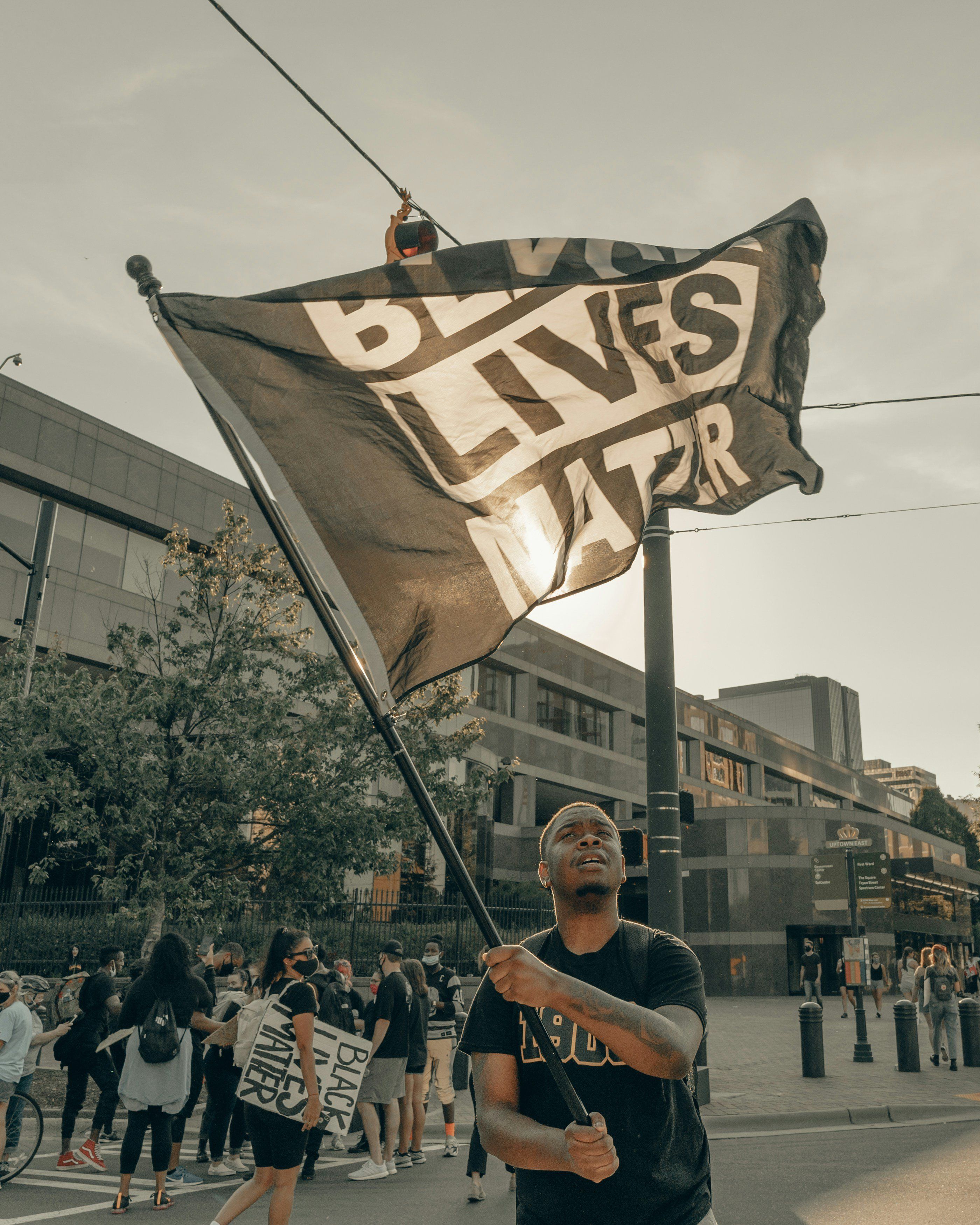 Black guy holding a large "Black Lives Matter" flag at a protest with buildings in the background.