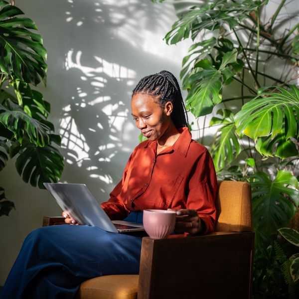 Giggling black woman using laptop. Cheerful laughing african american girl in chair among plants, drinking coffee from mug looking at computer screen, watching video movie series online on internet.