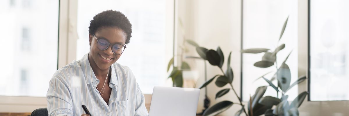 Inspired smiling female student, freelancer, office employee in glasses using the laptop for remote work, making research and taking notes, african-american business woman in modern coworking space