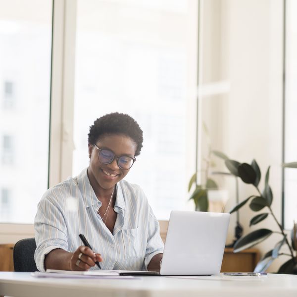 Inspired smiling female student, freelancer, office employee in glasses using the laptop for remote work, making research and taking notes, african-american business woman in modern coworking space