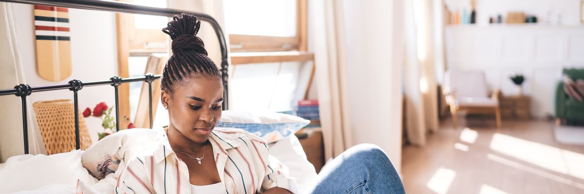 Stylish young Black woman sitting on bed, deeply immersed in reading book, with relaxed body language and casual clothes in sunlit minimalist bedroom