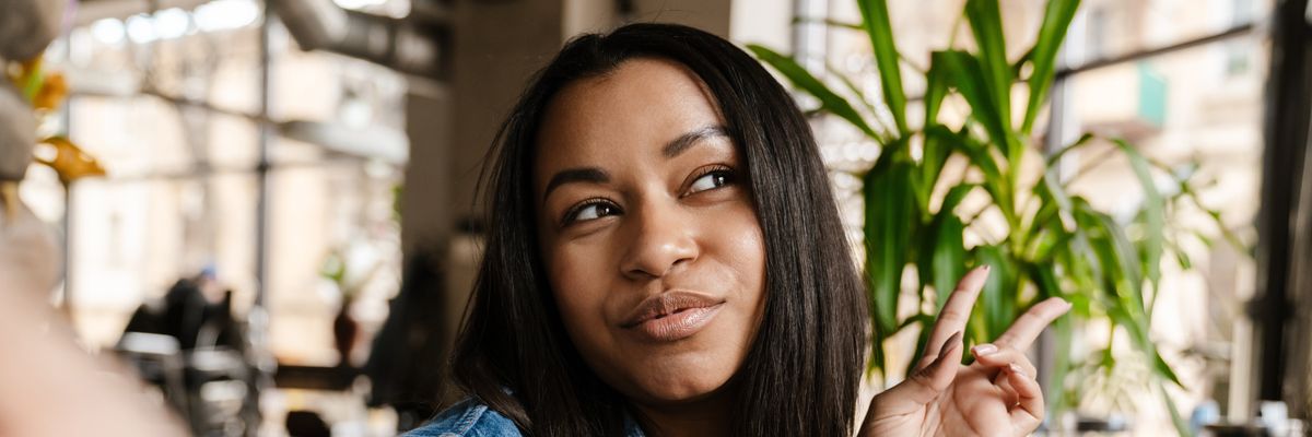 Black smiling woman gesturing peace sign and taking selfie photo while sitting in cafe