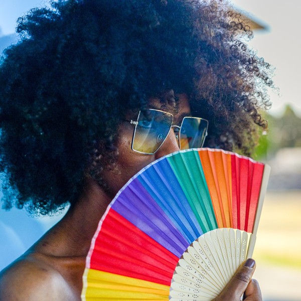 Young Black woman with afro hair and sunglasses cooling off with a colorful rainbow hand fan in summer heat