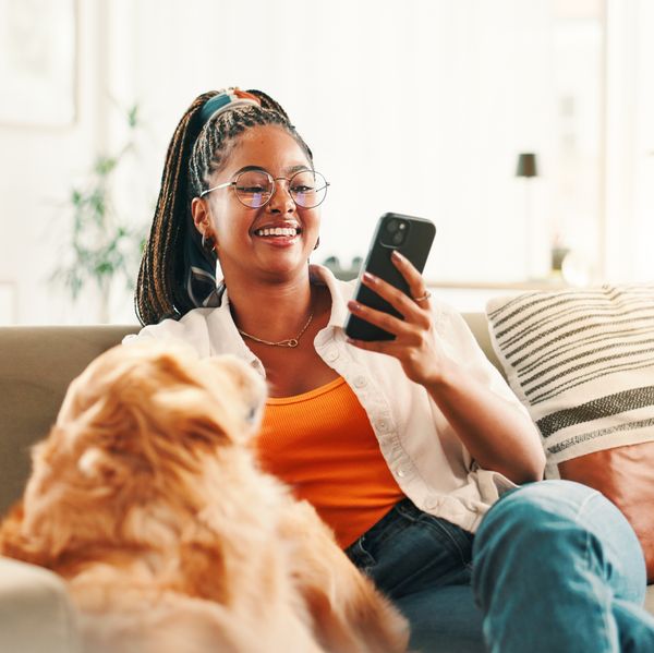 Black woman relaxing on sofa with dog while scrolling on smartphone