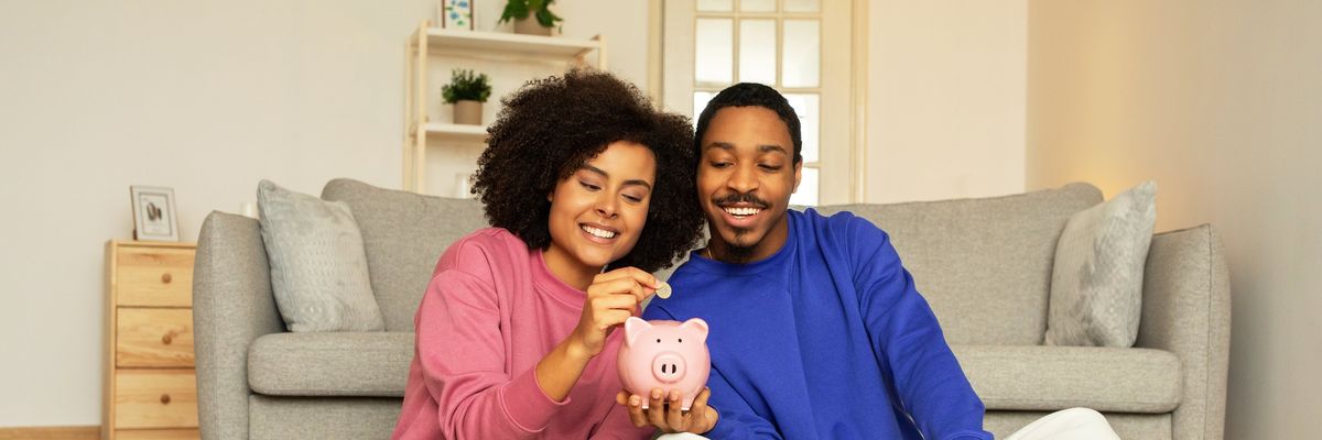 Young African American married couple happily posing with piggy bank, putting money savings sitting together at home. Concept of financial stability, smart money management and future wealth