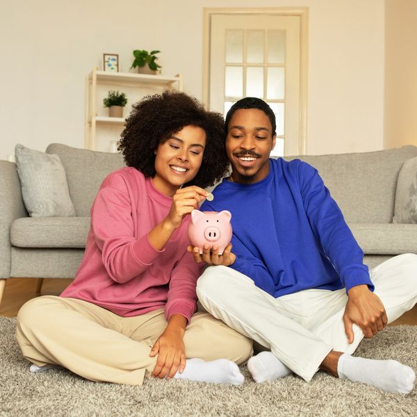 Young African American married couple happily posing with piggy bank, putting money savings sitting together at home. Concept of financial stability, smart money management and future wealth