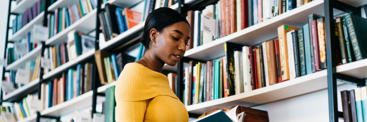 Focused African American lady in casual clothes reading textbook while standing near bookcase and studying in library of contemporary university