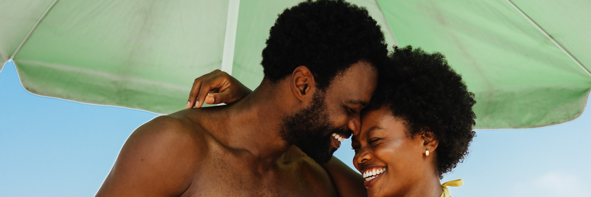 A loving couple shares a moment of affection during a baecation in Brazil. The tropical setting and clear skies highlight the joy and relaxation of their holiday, as they hold a coconut drink.