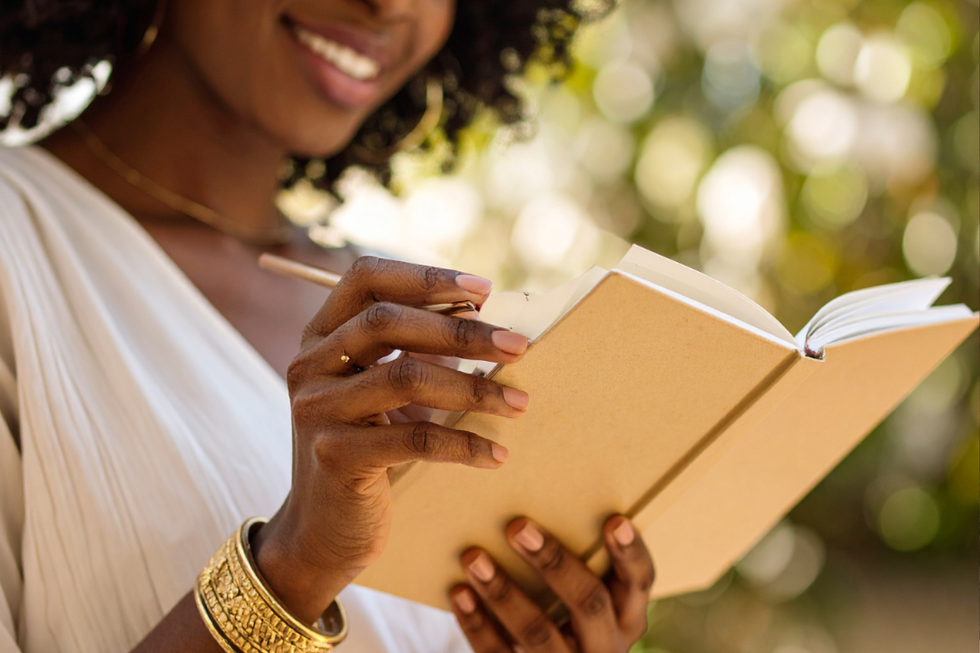 Woman writing in a notebook outdoors, smiling while holding a pencil.