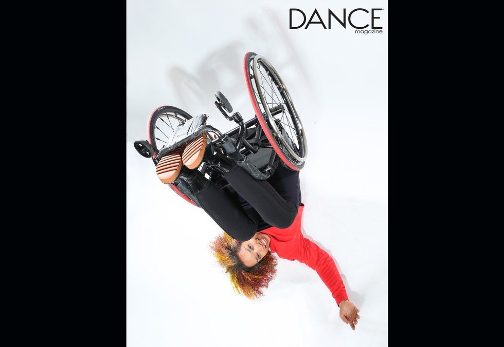 Alice Sheppard grins while lying on the floor, peeking out from under her wheelchair. She wears a bright red top that echoes the color of her wheels. Photo by Jayme Thornton.