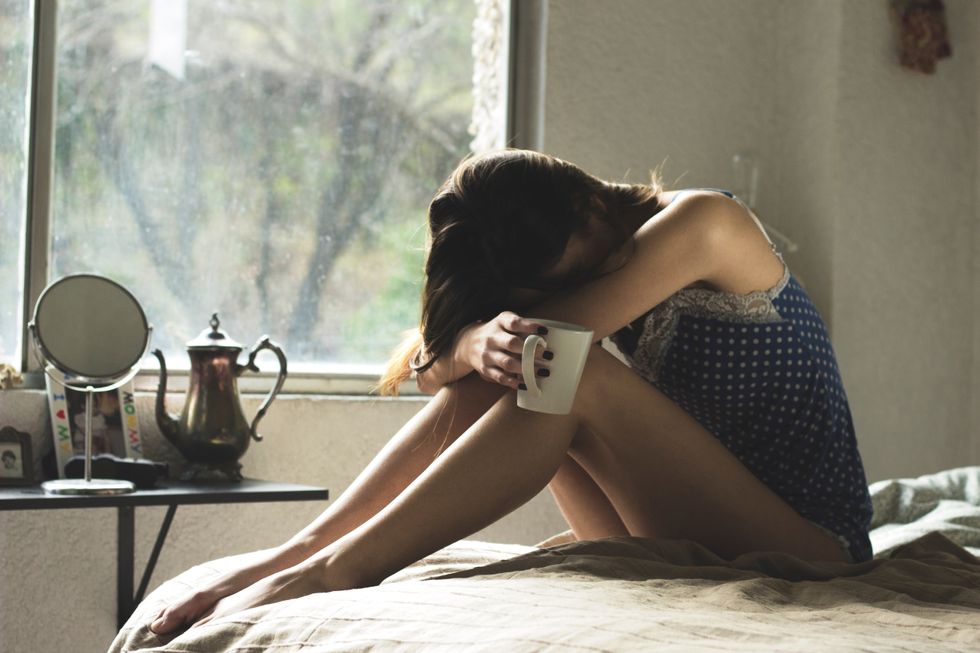 Girl sadly sitting on a bed with a mug in hand