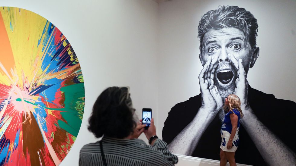 Visitors take a picture in front of a pictures of the British singer David Bowie and beside by one of the artworks (L) he owned named 'BEAUTIFUL, SHATTERING, SLASHING, VIOLENT, PINKY, HACKING, SPHINCTER PAINTING' by British artist Damien Hirst. (DANIEL LE