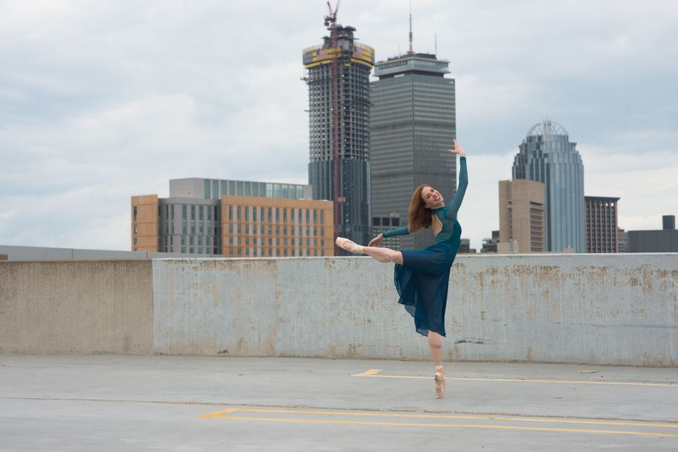 Schwan, in a blue long-sleeved leotard, skirt and pointe shoes, stands in back attitude on an urban roof, looking back at the camera and smiling.