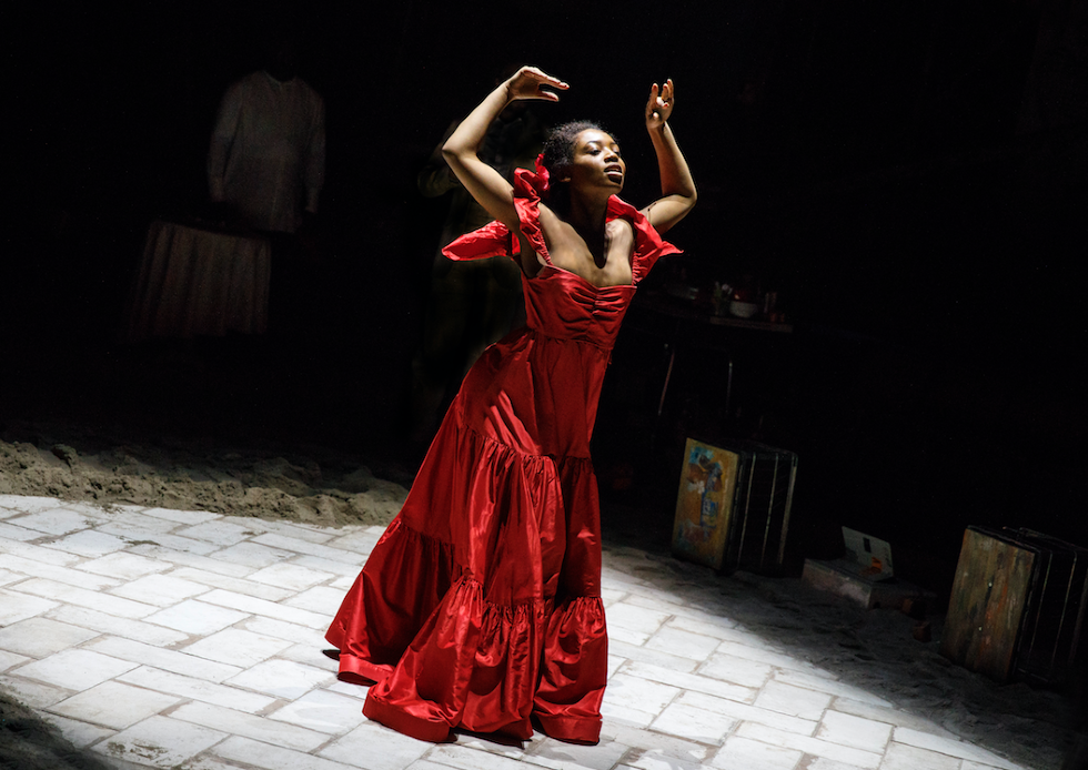 An actress in a red dress leans forward onstage, her hands above her head
