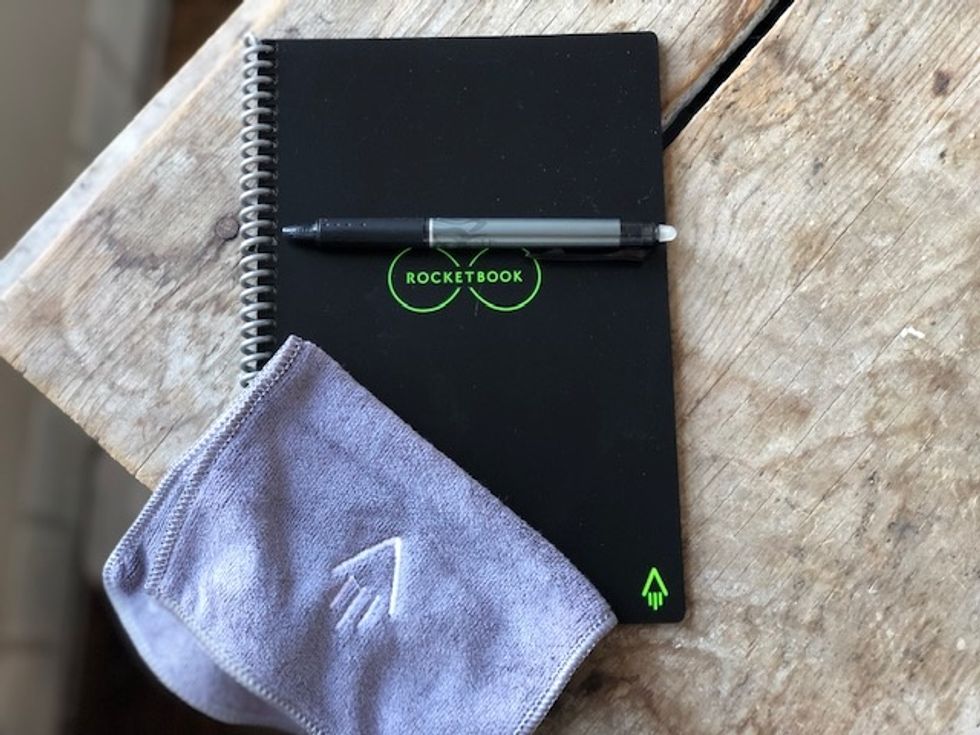 A photo of the Rocketbook Everlast reusable digital notebook in black on a wooden table, with a pen across it and a cloth sitting in the left corner