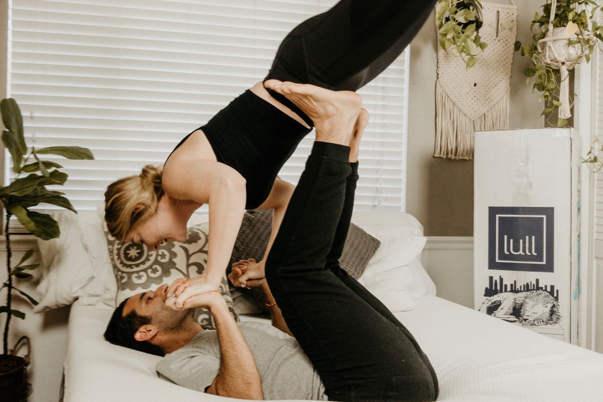 A man and woman in yoga clothes do acrobatics on a white bed