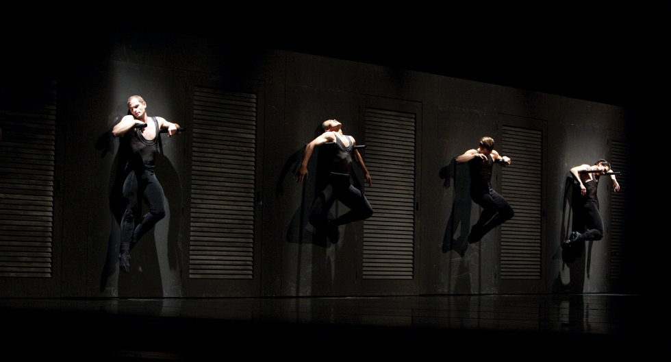 Four male dancers hang from a wall, all in black