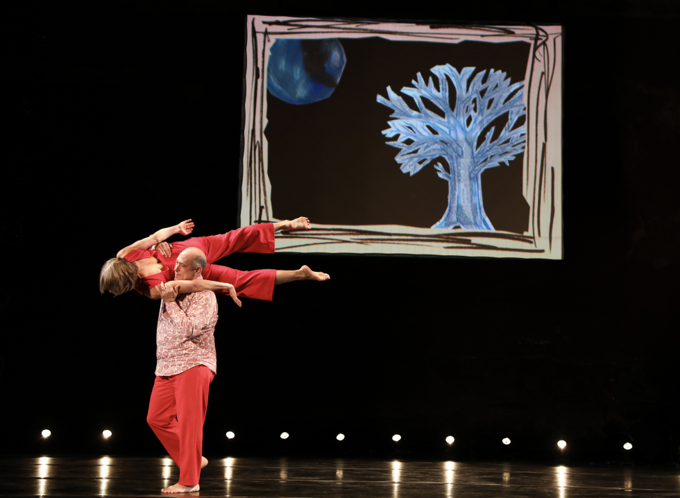 David Dorfman walks across a stage carrying a woman on his shoulders