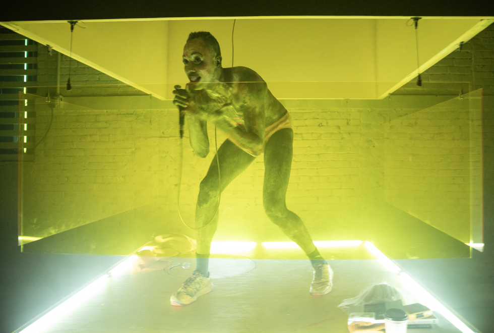Raja Feather Kelly in a yellow light holds a microphone and crouches