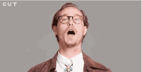 Person in glasses and brown jacket singing with mouth open, gray background.