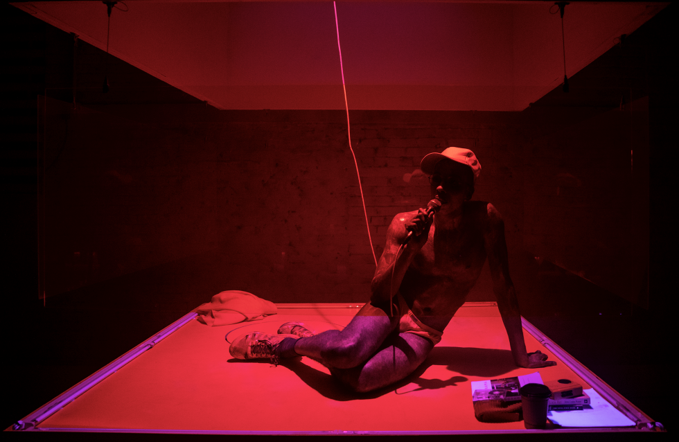 Raja Feather Kelly sits on a stage coated in red light, with a baseball cap and a microphone