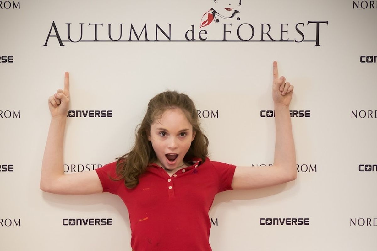 Girl in red shirt pointing up at "Autumn de Forest" sign with surprise.