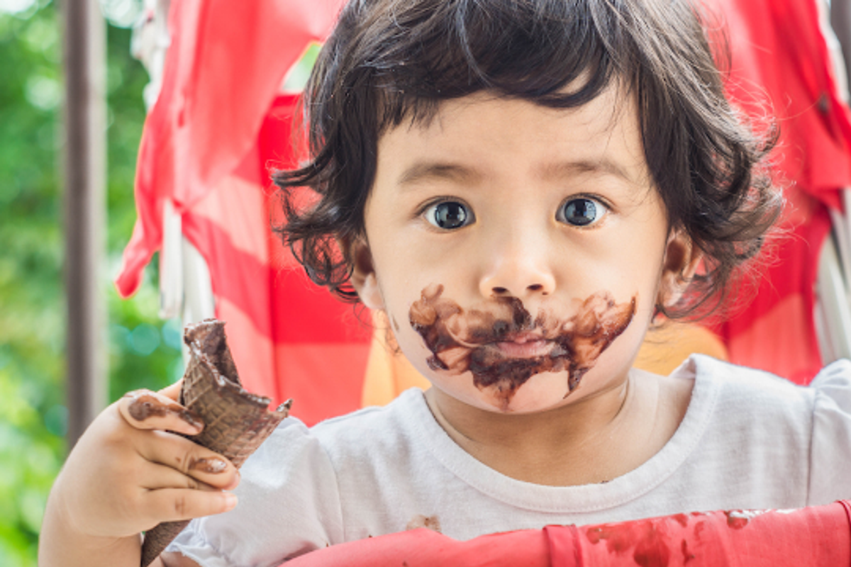 Child with chocolate-covered face holding an ice cream cone.