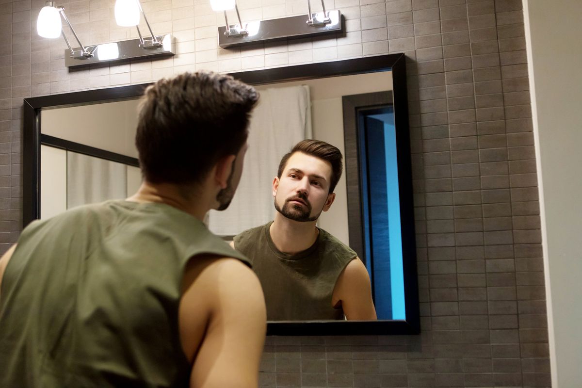 A brown-haired man in a green shirt with a sculpted beard stares at his mirror reflection