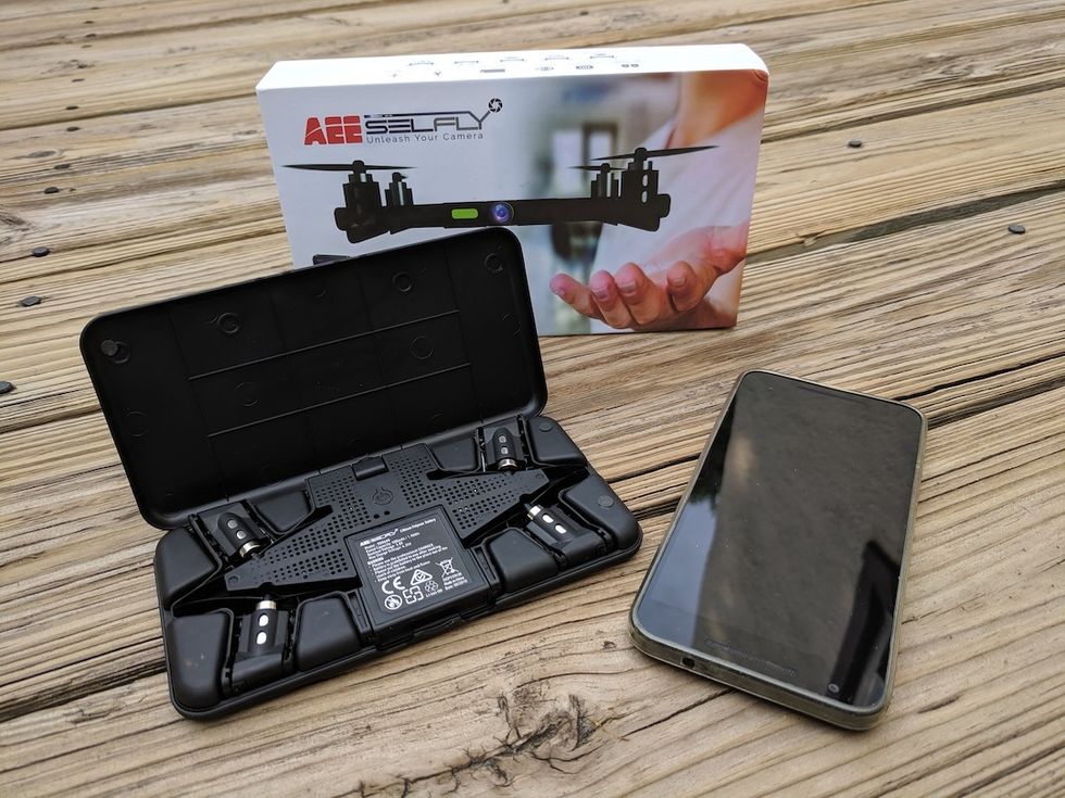 A drone folded into a black carrying case, next to a smartphone and both on top of a wooden picnic table