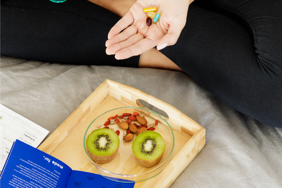 woman with vitamins in hand and fruit and nuts on plate while sitting in bed