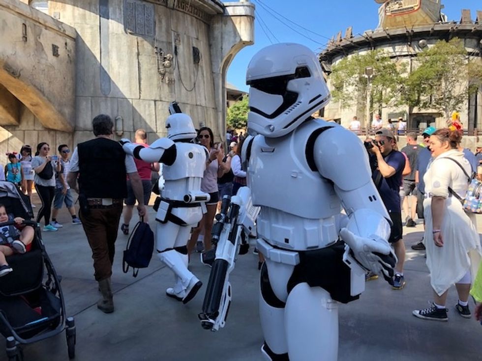 A Stormtrooper, standing, dressed in a white helmet and armor as people mill around