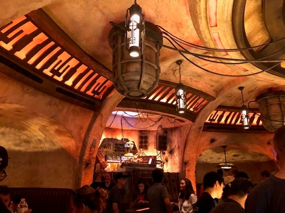 A robot in the background of a room lit in orange lights, with hoses draping down from the ceiling