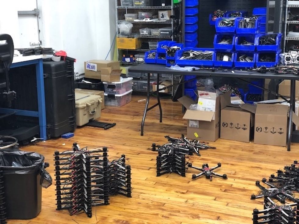 Drones stacked in piles inside a workspace with tables, blue bins and cardboard boxes