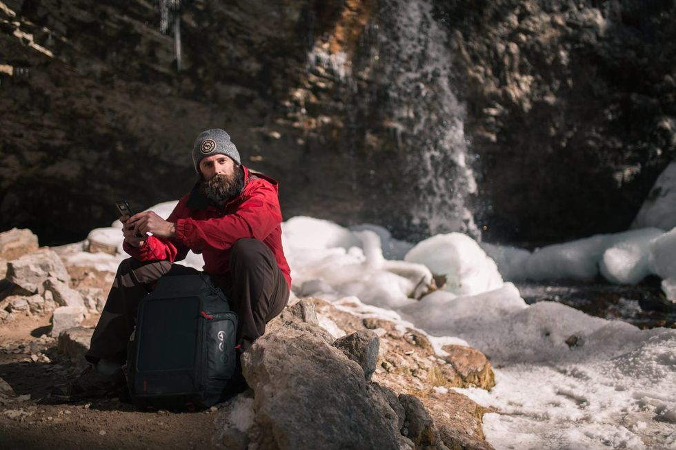 A figure with a beard and red jacket sitting next to a backpack near a river with rapids