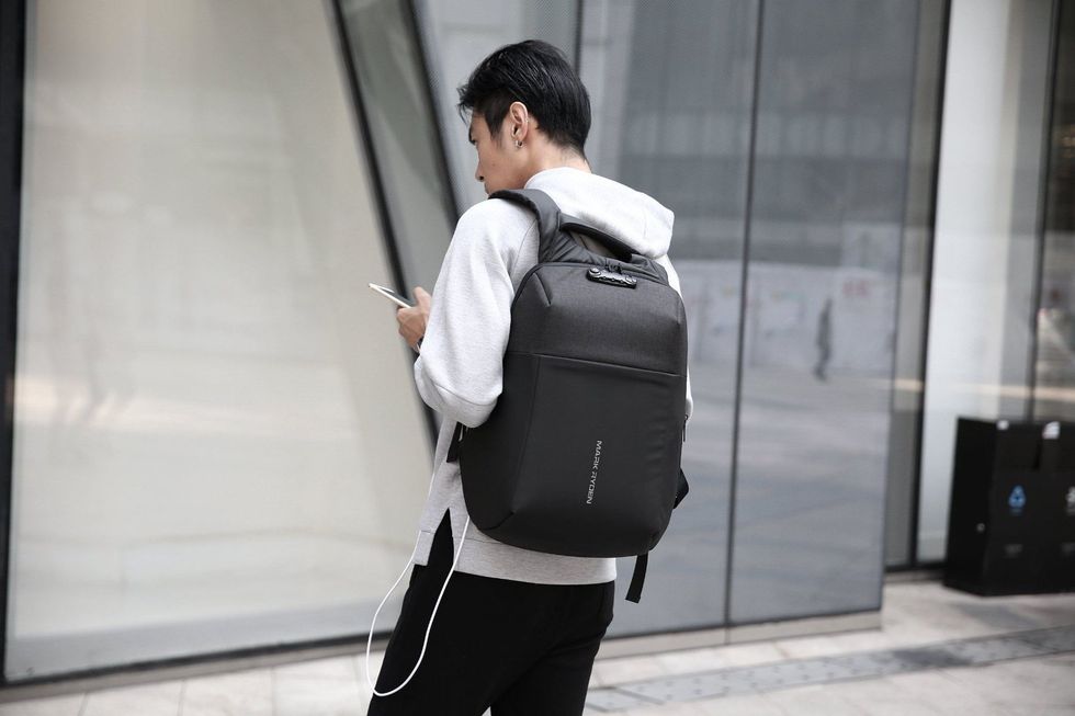 A figure with short hair wearing a black backpack and holding a smartphone with a charging wire leading back to the bag