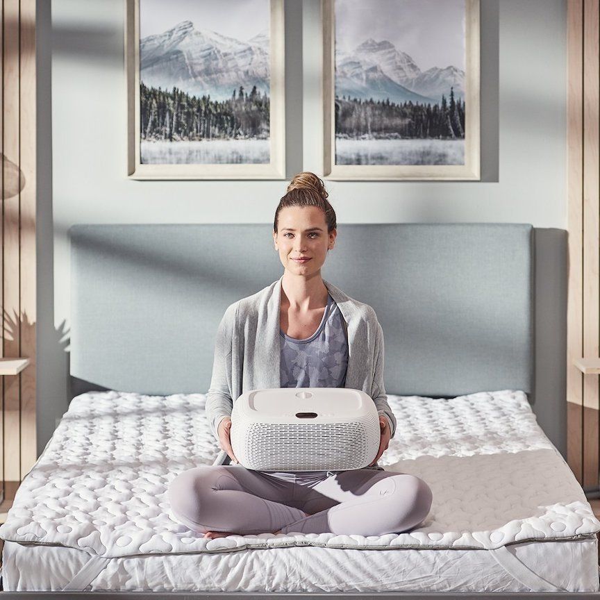 A smiling figure with hair tied up in a bun, seated on an unmade bed with a fan in their hands.