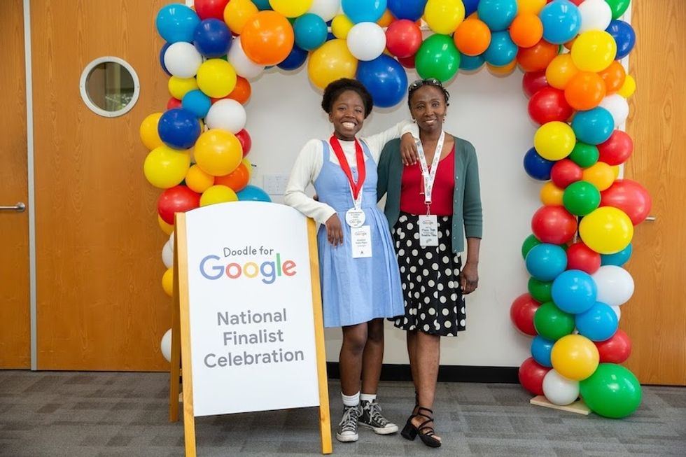 Two figures, one in a blue dress and one in a black and white polka dot skirt and red shirt smiling with their arms around each other, under a banner of balloons